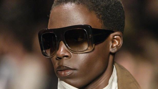 Person mit großer, dunkler Sonnenbrille, kurzem Haar, beigem Mantel und weißem, hochgeschlossenem Oberteil bei der Pariser Fashion Week