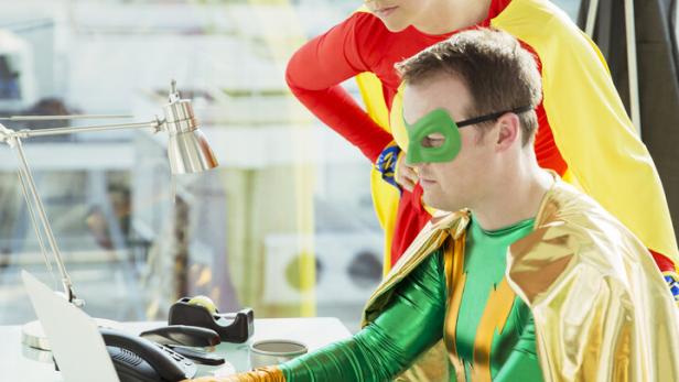 Zwei Erwachsene in Superheldenkostümen arbeiten gemeinsam an einem Schreibtisch mit Laptop in einem modernen Büro.