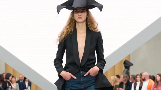 Eine Frau trägt einen tief ausgeschnittenen schwarzen Blazer, einen Jeansrock und einen auffälligen schwarzen Hut auf einem Laufsteg.
