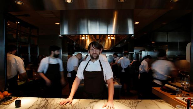 Starkoch und Noma-Chef René Redzepi steht an einer Marmorarbeitsfläche, während mehrere Köche im Hintergrund arbeiten.