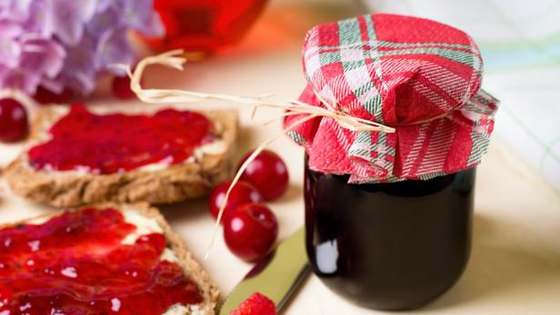Ein Marmeladenglas mit rot-kariertem Deckel steht neben Kirschen und Brotscheiben mit rotem Aufstrich.