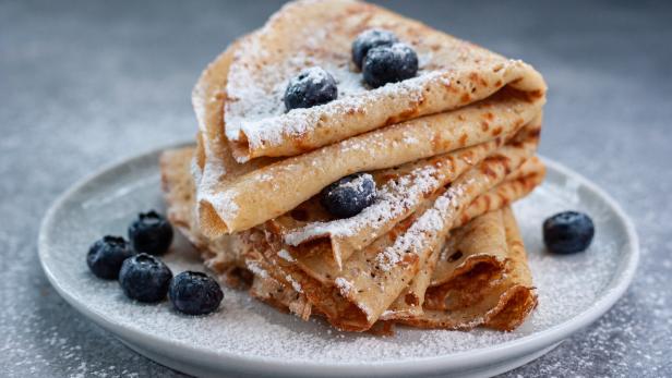 Mehrere zusammengefaltete Crêpes mit Puderzucker bestreut und mit frischen Heidelbeeren garniert auf einem Teller.