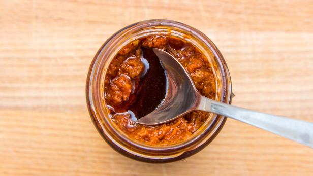 Ein Glas mit rotem Pesto und einem Löffel steht auf einem Holztisch.