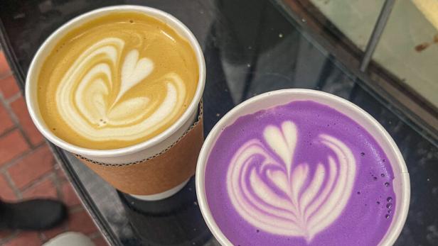 Zwei Becher mit Latte Art, einer mit gelbem und einer mit lila Getränk, stehen nebeneinander auf einem Tisch.