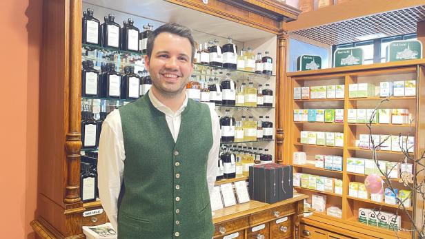 Ein Mann mit grünem Gilet steht lächelnd in einer traditionellen Apotheke vor Regalen mit Flaschen und Produkten.