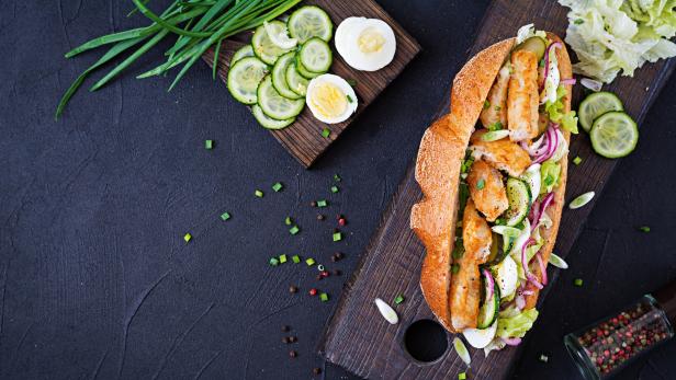 Ein belegtes Baguette mit paniertem Hähnchen, Gurken, Eiern, Salat und Zwiebeln auf einem Holzbrett, daneben frische Zutaten.