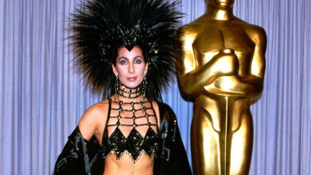 Cher posiert in einem extravaganten, schwarzen Outfit mit Federschmuck neben einer goldenen Oscar-Statue.