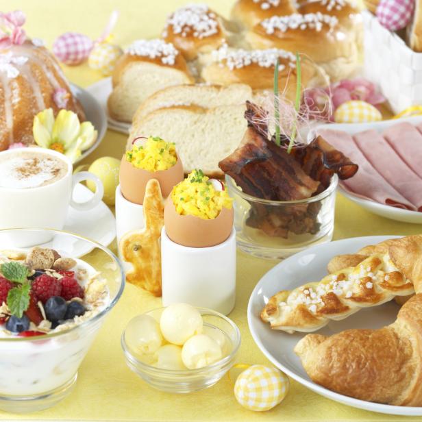 Ein reichhaltiges Osterfrühstück mit Kuchen, Croissants, Eiern, Müsli und Aufschnitt auf einem gelben Tisch.