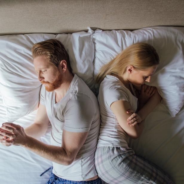 Young couple lying in bed back to back, man using phone and woman frowning