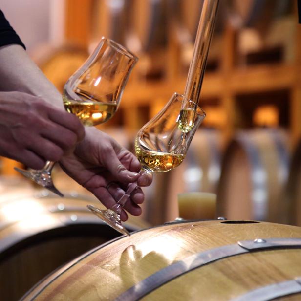Zwei Hände halten Gläser mit bernsteinfarbenem Whisky über einem Holzfass.