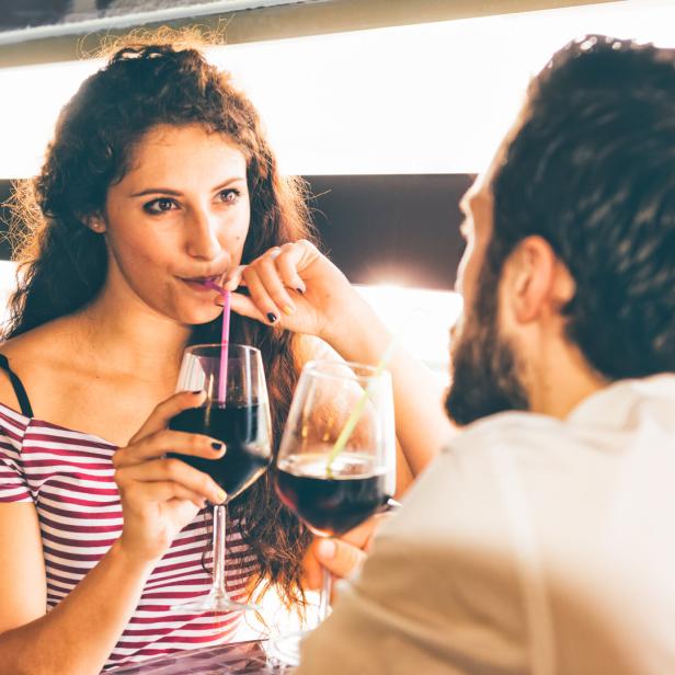 Eine Frau mit lockigem Haar und ein bärtiger Mann sitzen sich gegenüber und trinken ein Getränk aus Gläsern mit Strohhalmen.