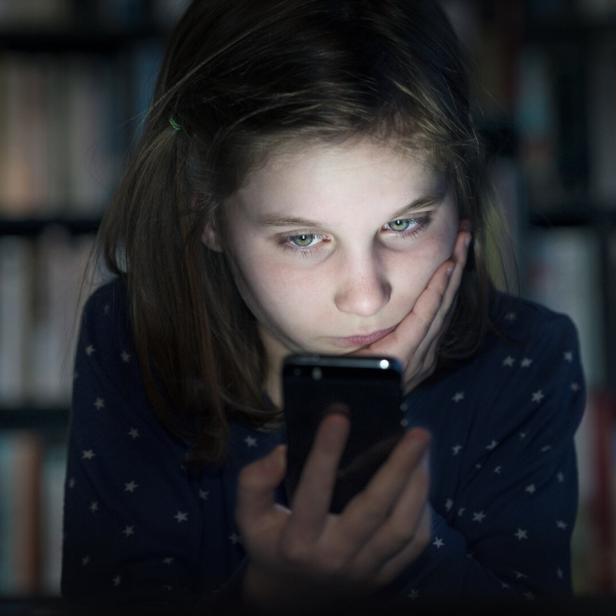 Ein Mädchen schaut mit ernstem Blick auf ihr Smartphone, im Hintergrund ein Bücherregal.