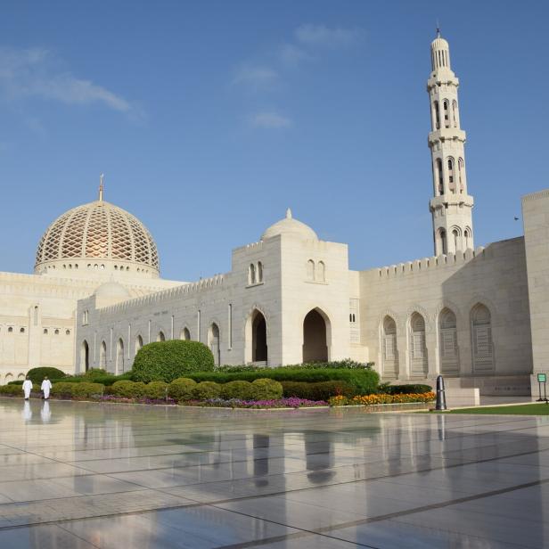 Weiße Moschee mit Kuppel und Minarett vor blauem Himmel, davor spiegelnder Platz und Blumenbeete.