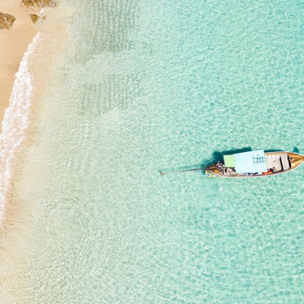 View from above, stunning aerial view of two people walking on a beautiful tropical beach with white sand, turquoise clear water and a traditional long-tail boat. Banana beach, Phuket, Thailand.