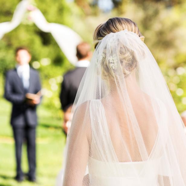 Eine Braut mit Schleier steht im Freien vor einem Traubogen, während Gäste und ein Redner warten.