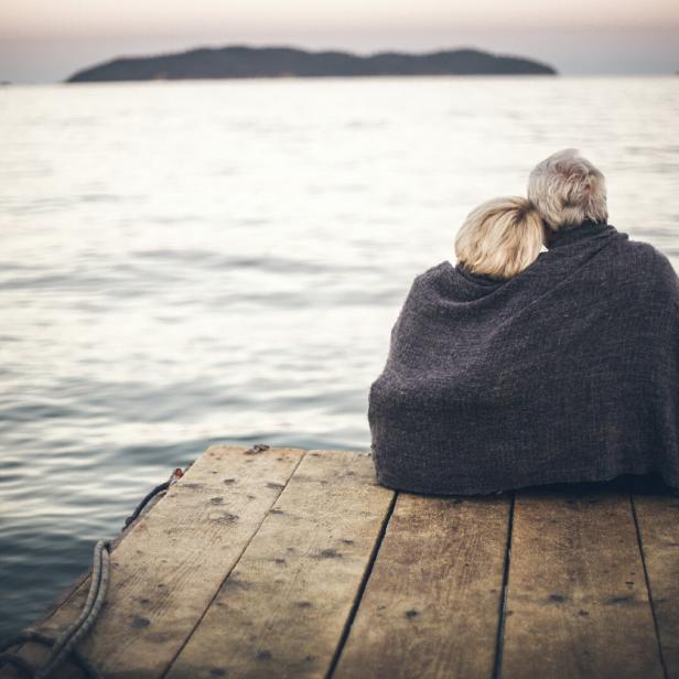 Ein älteres Paar sitzt gemeinsam unter einer Decke auf einem Holzsteg am Meer und blickt auf das Wasser.