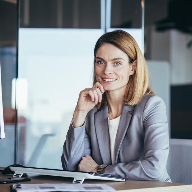 Happy and successful business woman boss working at computer in modern office looking at camera and smiling