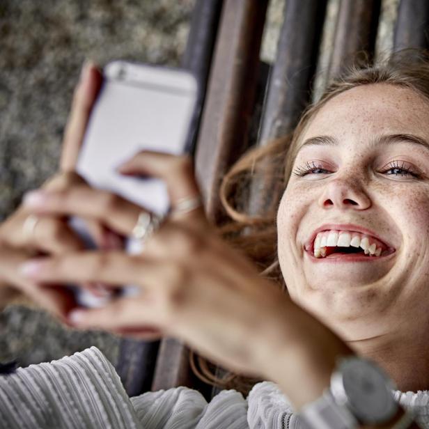 Laughing young woman lying on a bench using cell phone - Stock-Fotografie