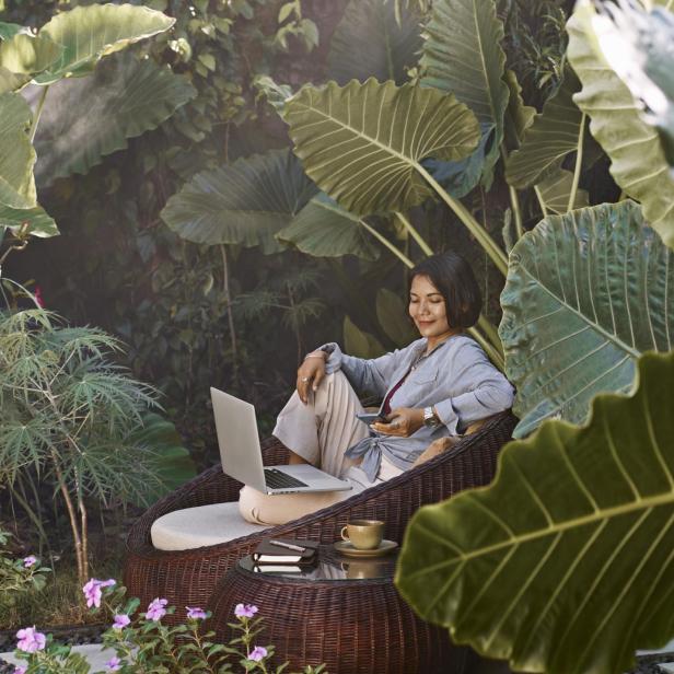 Asian woman working from home sitting in garden furniture surrounded by tropical plants, using laptop computer and mobile phone - Stock-Fotografie