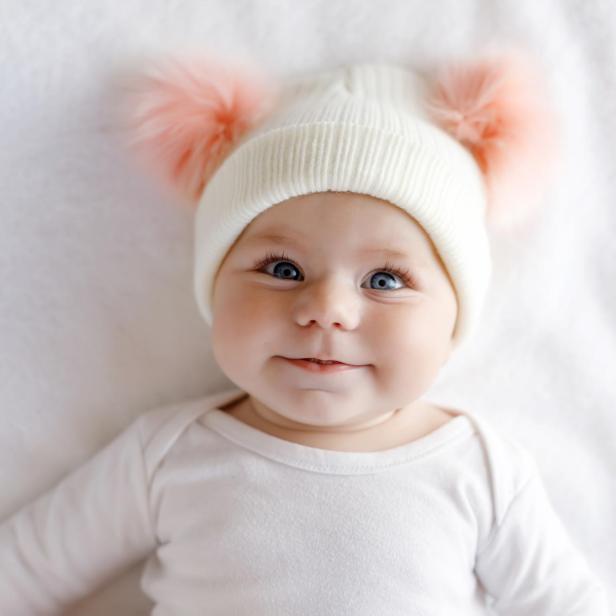Süßes entzückendes Babykind mit warmweißem und rosa Hut mit süßen Bobbles. Glückliches kleines Mädchen auf weißem Hintergrund und Blick in die Kamera. Nahaufnahme für Weihnachtsurlaub und Familienkonzept.