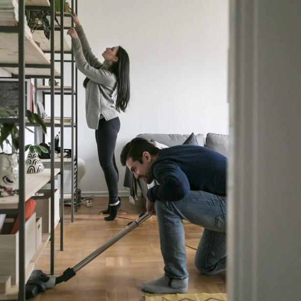 Man vacuuming floor while woman cleaning shelf at home - Stock-Fotografie