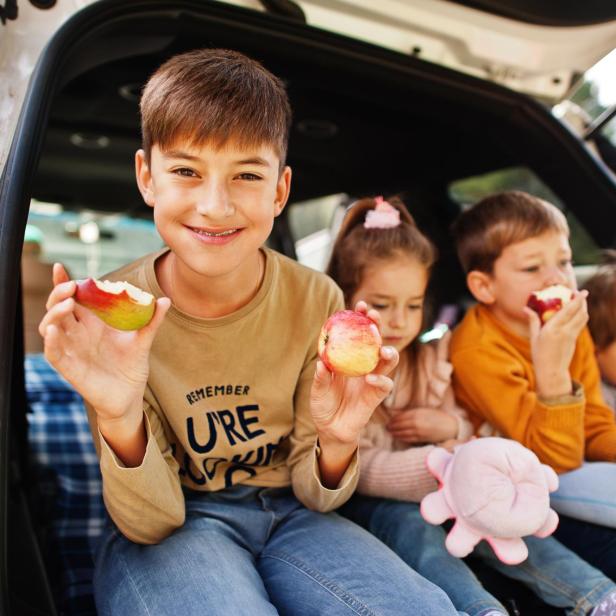 Familie mit vier Kindern isst Äpfel im Fahrzeuginnenraum. Kinder sitzen im Kofferraum. Anreise mit dem Auto in die Berge, Atmosphärenkonzept. - Stock-Fotografie