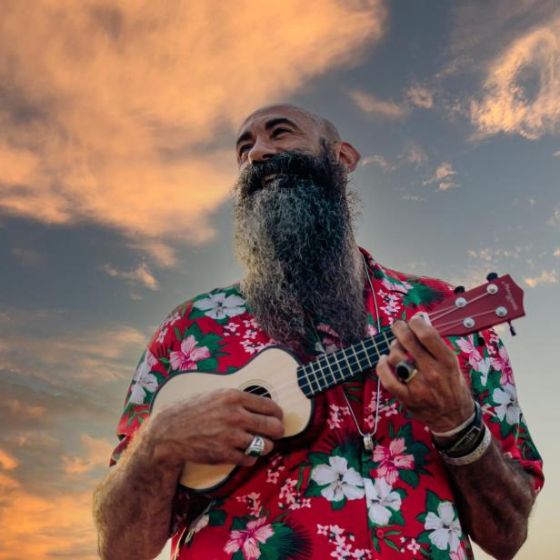 Rasierter Kopf reifer Mann mit langem Bart gekleidet mit Hawaiihemd spielt Ukulele mit einem dramatischen Himmel im Hintergrund - Stock-Fotografie