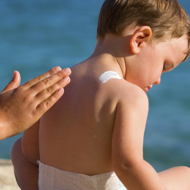 Ein Kind am Wasser bekommt Sonnencreme auf die Schulter aufgetragen.