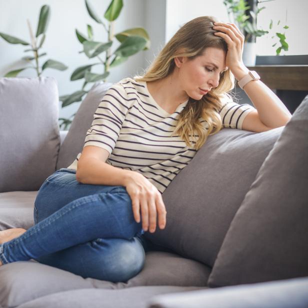 Eine Frau mit blonden Haaren sitzt nachdenklich auf einem grauen Sofa.