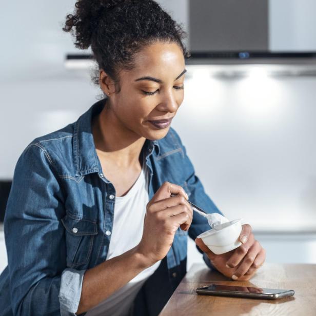 Woman eating yogurt sitting in kitchen at home - Stock-Fotografie