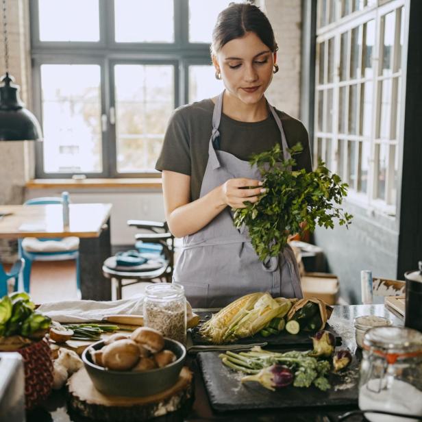Female chef wearing apron doing quality check of cilantro standing in studio kitchen - Stock-Fotografie