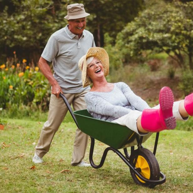 Älterer Mann schiebt glückliche Frau in der Schubkarre. Sie streckt die Füße in pinken Gummistiefeln lachend in die Luft.