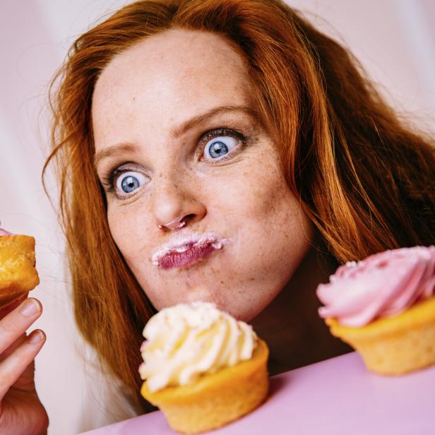 Eine rothaarige Frau mit Zuckerguss im Gesicht hält einen Cupcake und blickt mit großen Augen auf weitere Cupcakes.