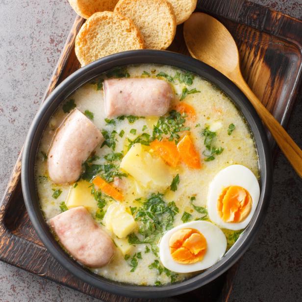 Polnische Suppe Zurek mit Weißwurst, Eiern, Kartoffeln und Karotten in einer schwarzen Schüssel, daneben ein Holzlöffel und Brot.