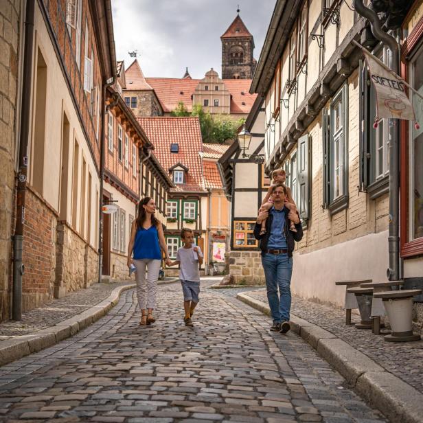Familie mit Kindern spaziert in mittelalterlicher Stadt in einer Gasse mit Kopfsteinpflaster, im Hintergrund sieht man eine Burg