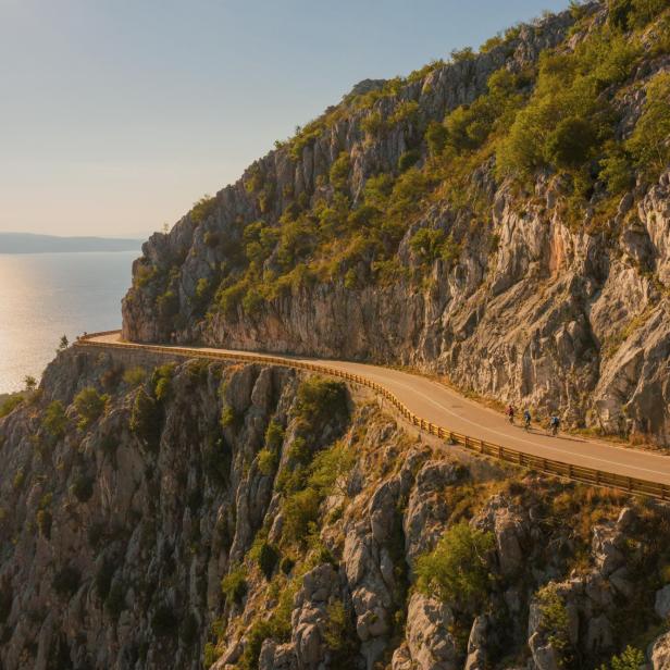 Eine Küstenstraße schlängelt sich an einer Klippe entlang, während Radfahrer die Aussicht genießen.