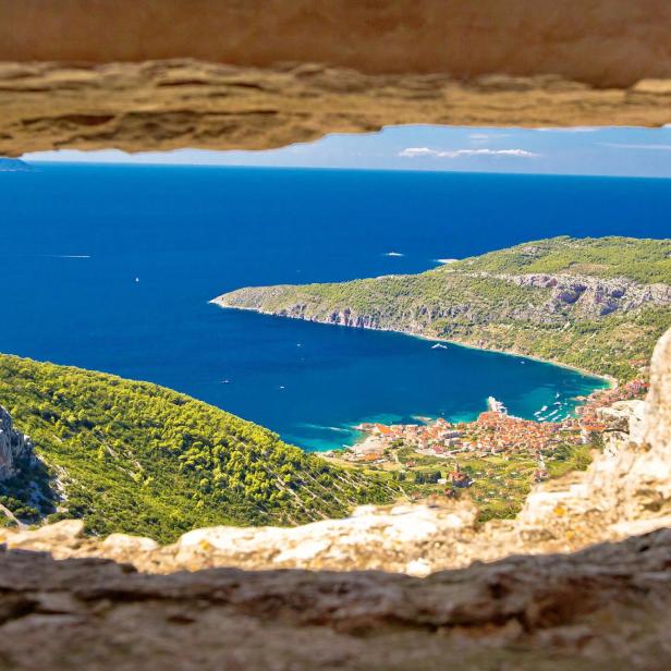 Blick durch eine steinerne Fensteröffnung auf eine Küstenstadt und das Meer.
