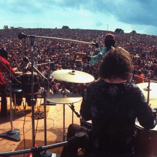 Eine Band auf der Bühne beim Woodstock Festival 1969. Von der Bühne blicken die Musiker inmitten eine Menschenmasse