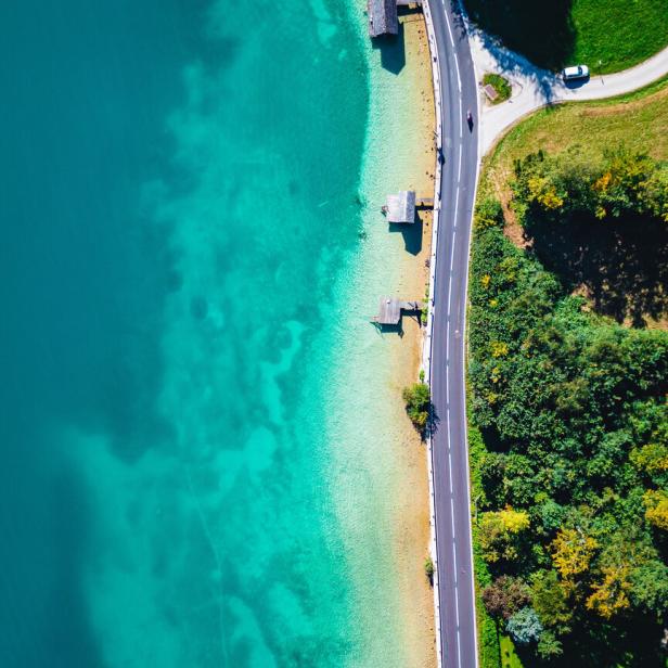 Eine Drohnenaufnahme vom Attersee: Eine Wiese mit Bäumen, dann eine Straße am Ufer und daran grenzt der klare Attersee in verschiedenen Blautönen.