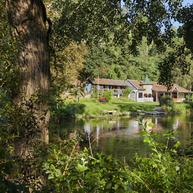 Grün-braunes Wasser, Bäume, eine Liegewiese und im Hintergrund ein Holzgebäude, das einst das Flussbad beherbergte