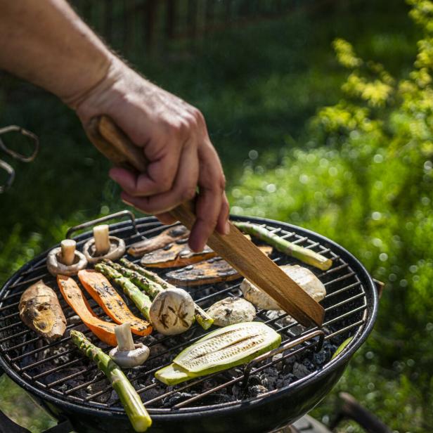 Kleiner Griller, auf dem Rost liegen Zucchini, Champignons, Spargel und Melanzani, ein Mann wendet das Gemüse mit einer Grillzange