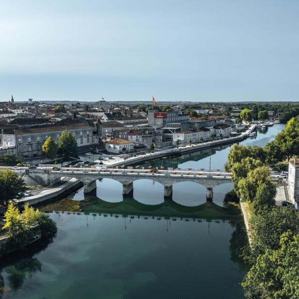 Eine Brücke über dem Fluss Charente. Am Ufer stehen Teile der Stadtmauer, ein großes Gebäude des Cognac-Herstellers Hennessey, im Hintergrund erhebt sich die Kirche.