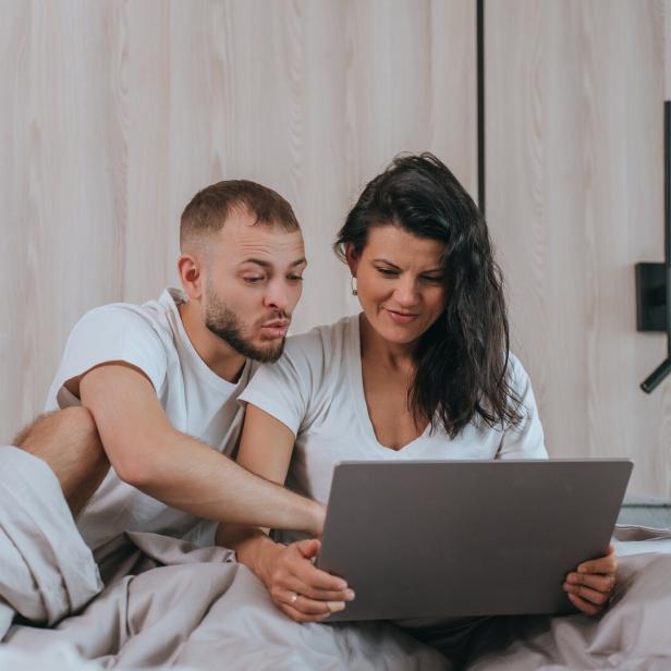 Ein Paar sitzt im Bett und schaut auf einen Laptop. 