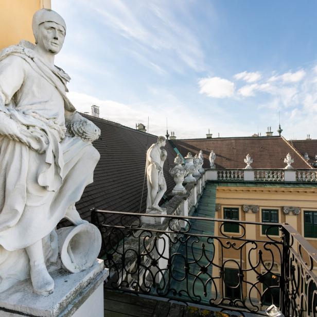 Steinfiguren schauen beim Balkon unterhalb der großen Uhr von oben auf den Ehrenhof des Schlosses Schönbrunn herab. Vor dem Balkon befindet sich ein Schmiedeisengitter, vor dem Dach eine steinerne Brüstung