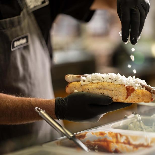 Eine Bosna entsteht: Hände in Handschuhen halten ein Baguette: Darin liegt eine Bartwurst, die Hand streut Zwiebeln darauf.