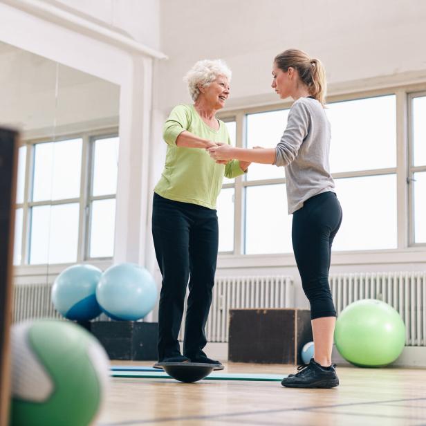 Eine Trainerin hilft einer Seniorin auf dem Balance-Board