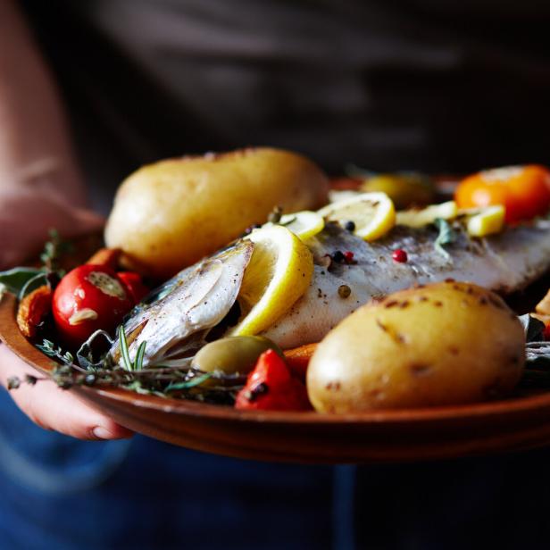 Ein ganzer gebratener Fisch mit Zitronenscheiben, Kartoffeln, Tomaten und Kräutern auf einem Teller.