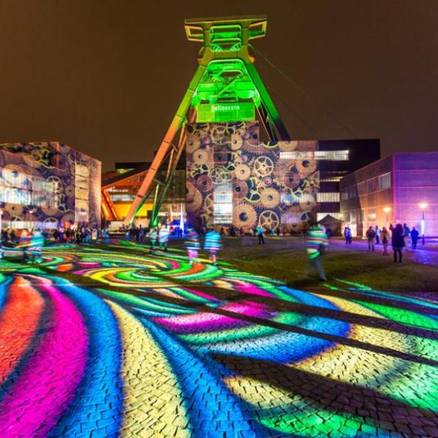 Die Zeche Zollverein in Essen ist nachts mit bunten Lichtern und Zahnrädern illuminiert.