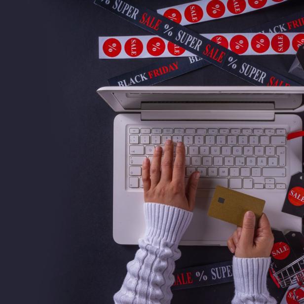 Person in weissem Pullover tippt auf einem Laptop und hält eine Kreditkarte in der anderen Hand; rundherum liegen Geschenkschachteln, ein kleiner Einkaufswagen und Bänder mit Aufschriften wie "Black Friday", "Sale" und "Super Sale" auf dunklem Hintergrund.