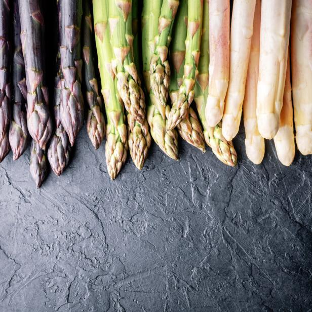 Drei Sorten Spargel, lila, grün und weiß, liegen nebeneinander auf dunklem Untergrund.
